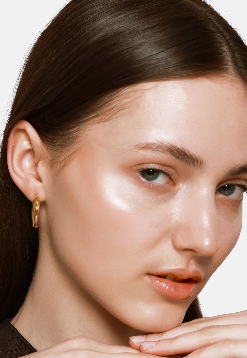 Boucle d'oreille en or, forme ronde, surface brillante. Texture de peau lisse avec un éclat naturel, cheveux châtain clair et couleur de lèvres neutre.