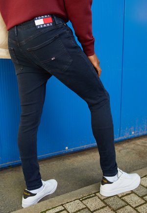 Person trägt dunkle Skinny Jeans mit Tommy Jeans-Logo und weißen Sneakers, steht auf einem Bürgersteig vor einer blauen Wand.