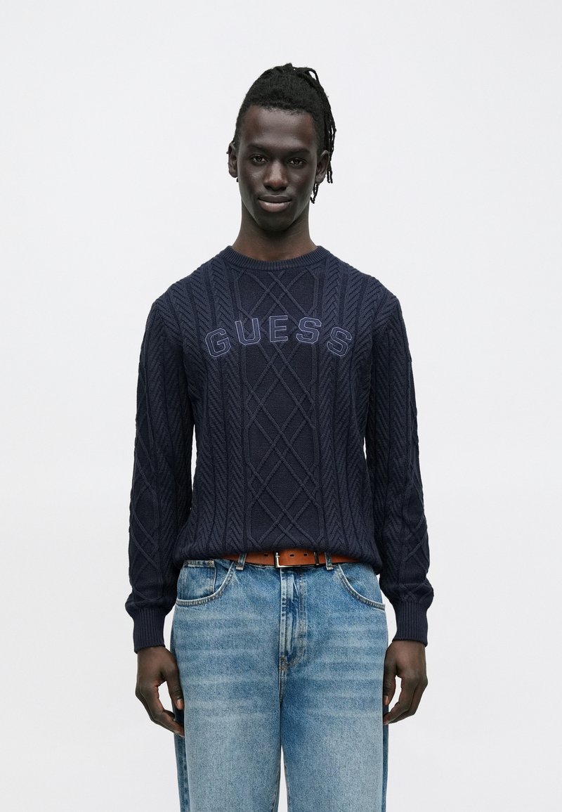 Mann mit dunklen, geflochtenen Haaren, der einen marineblauen Strickpullover mit "GUESS"-Schriftzug und hellblaue Jeans vor weißem Hintergrund trägt.