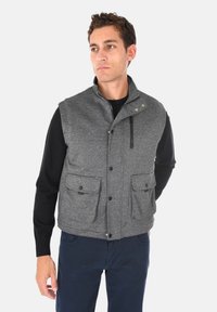 Gilet grigio in misto lana con colletto alto, bottoni frontali, due ampie tasche a toppa e tasca sul petto con zip. Camicia nera a maniche lunghe sotto.