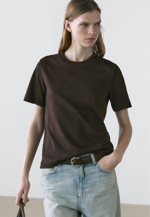 Jonge vrouw die een donkerbruin shirt met korte mouwen draagt, ingestopt in lichtblauwe jeans met een bruine riem, en een donkere tas in haar linkerhand vasthoudt.