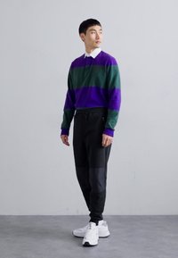 Camisa polo de manga longa em riscas verdes e roxas, com colarinho branco. Combinada com calças pretas ajustadas e ténis brancos. Detalhe de cintura ajustável.