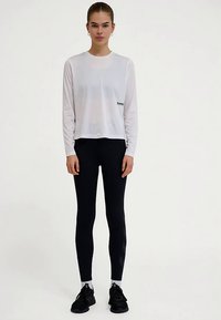 Witte longsleeve met een afgeronde halslijn en logo bij de zoom, gecombineerd met zwarte leggings met een getextureerd zijpatroon en zwarte sneakers.
