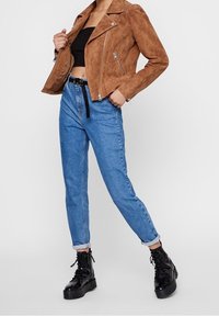 Veste en daim marron avec des zips argentés, top court noir, jean taille haute bleu clair, et bottes à lacets noires à plateforme.
