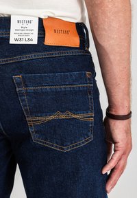 Ciemnoniebieskie dżinsy z denimu o średnim stanie, prostych nogawkach i pomarańczową skórzaną metką. Charakteryzują się kontrastowym żółtym przeszyciem i wzorem kieszeni.