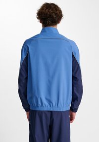 Blå och marinblå idrottsjacka med stående krage, elastiska manschetter och midja. Har kontrasterande färger och en slät textur.