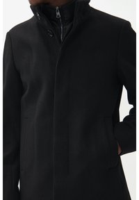 Mantello in misto lana nero con colletto alto, chiusura con zip, due tasche laterali e dettagli in bottone, caratterizzato da un tessuto elegante e testurizzato.