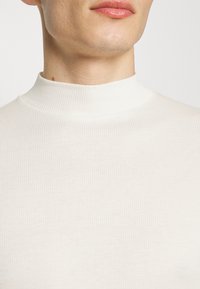 Top de color crema con cuello alto y canalé, que presenta un tejido liso con textura y un diseño sin costuras alrededor del cuello y los hombros.