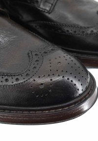 Scarpe di pelle nera con punta arrotondata, dettagli brogue, leggero lucido e una welt in pelle marrone a contrasto. Superficie texture con perforazioni.