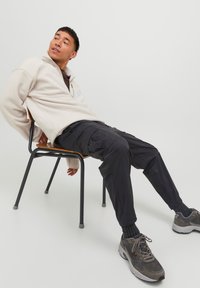 Pullover in pile crema con mezza zip, pantaloni cargo neri con tasche, sneakers grigie. Il pullover ha una spalla scesa e i pantaloni sono affusolati.
