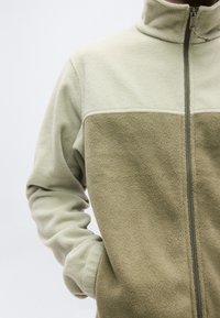 Ζακέτα από φλις με φερμουάρ, με ανοιχτό πράσινο πάνω μέρος και σκουρότερο πράσινο κάτω μέρος, υφή υφάσματος και πλαϊνές τσέπες.