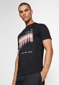 Črna bombažna majica s kratkimi rokavi, s belim logotipom "LACOSTE" in roza degradiranim besedilom. Zeleno vezeno krokodilovo okrasje nad logotipom.
