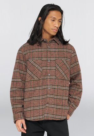 Homme aux longs cheveux noirs portant une chemise à carreaux marron et rouge boutonnée avec des poches, regardant vers le bas et sur le côté, debout devant un fond uni.