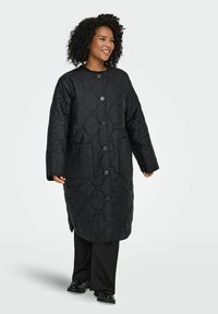 Juodas siuvinėtas ilgas paltas su apvalia apačia, didelėmis priekinėmis kišenėmis, užsegamas mygtukais priekyje ir lygia tekstūra.