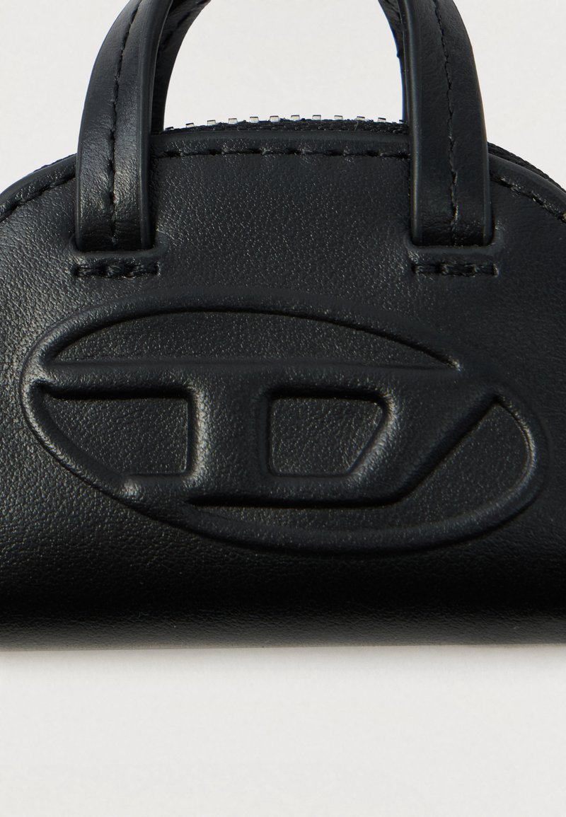 Gros plan sur un sac à main en cuir noir montrant un logo ovale en relief et des anses cousues sur fond uni.