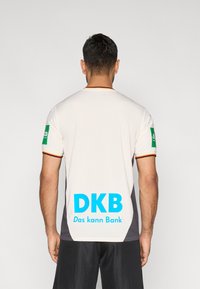 Balts un pelēks sporta krekls ar īsām piedurknēm, krāsainu apmali, lielu zilu "DKB" logotipu uz muguras un zaļiem sponsoru uzlīmiem.