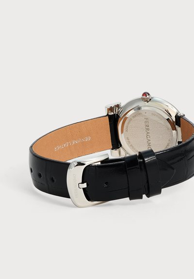 Μαύρο δερμάτινο ρολόι χειρός με ασημένια αγκράφα και στρογγυλή ασημένια πίσω όψη θήκης, χαραγμένη με την επωνυμία Ferragamo και "Genuine Leather" στο εσωτερικό του λουριού.