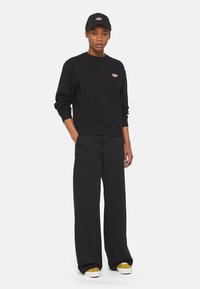 Felpa nera con collo rotondo e polsini a costine, abbinata a pantaloni neri a gamba larga. Un cappellino nero coordinato presenta un piccolo logo.