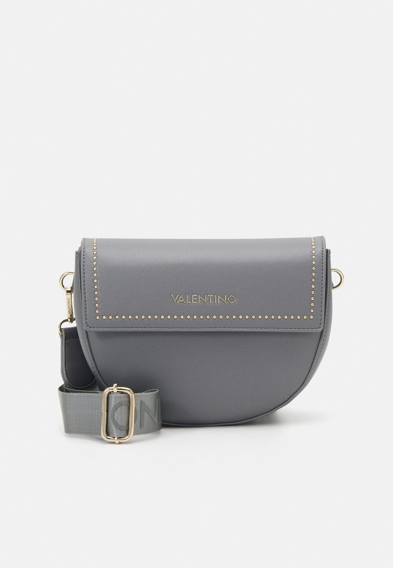 Šedá kožená crossbody taška s kulatým tvarem, zlatými hroty a logem. Obsahuje odnímatelný popruh s vyznačenou sponou.