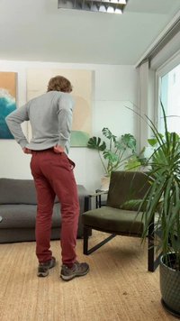 Calças cor de vinho e suéter cinza usados por uma pessoa vista por trás, em pé ao lado de uma poltrona verde e plantas de interior numa sala.