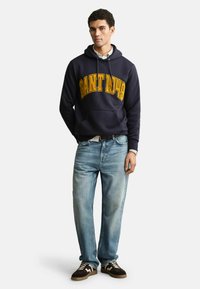 Mann trägt einen dunkelblauen Hoodie mit gelbem "GANT 1949"-Schriftzug, hellblaue Jeans, braune Sneaker und ein weißes Hemd darunter, steht vor weißem Hintergrund.