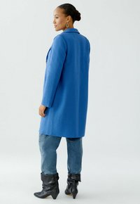 Blauwe wolmix jas met een gestructureerde afwerking, lange mouwen, een notch-kraag en twee knopen. Gedragen over lichtblauwe jeans en zwarte slouchy laarzen.