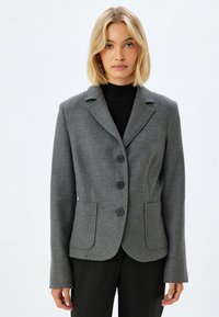Blazer grigio in misto lana con rever a punte, tre bottoni neri, due tasche frontali e vestibilità sartoriale. Maniche lunghe, tessuto liscio.