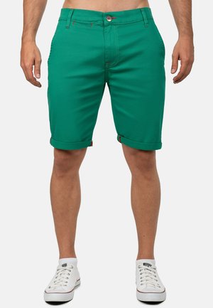 Shorts en coton verts avec un design ajusté, ourlet roulé et détails de couture rouges contrastants. Portés avec des baskets blanches.