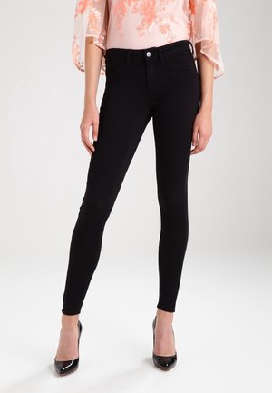 Kobieta nosząca czarne, wysokiej talii dopasowane rurki, skomponowane z różową, kwiatową koronkową bluzką i czarnymi lakierowanymi szpiczastymi szpilkami.