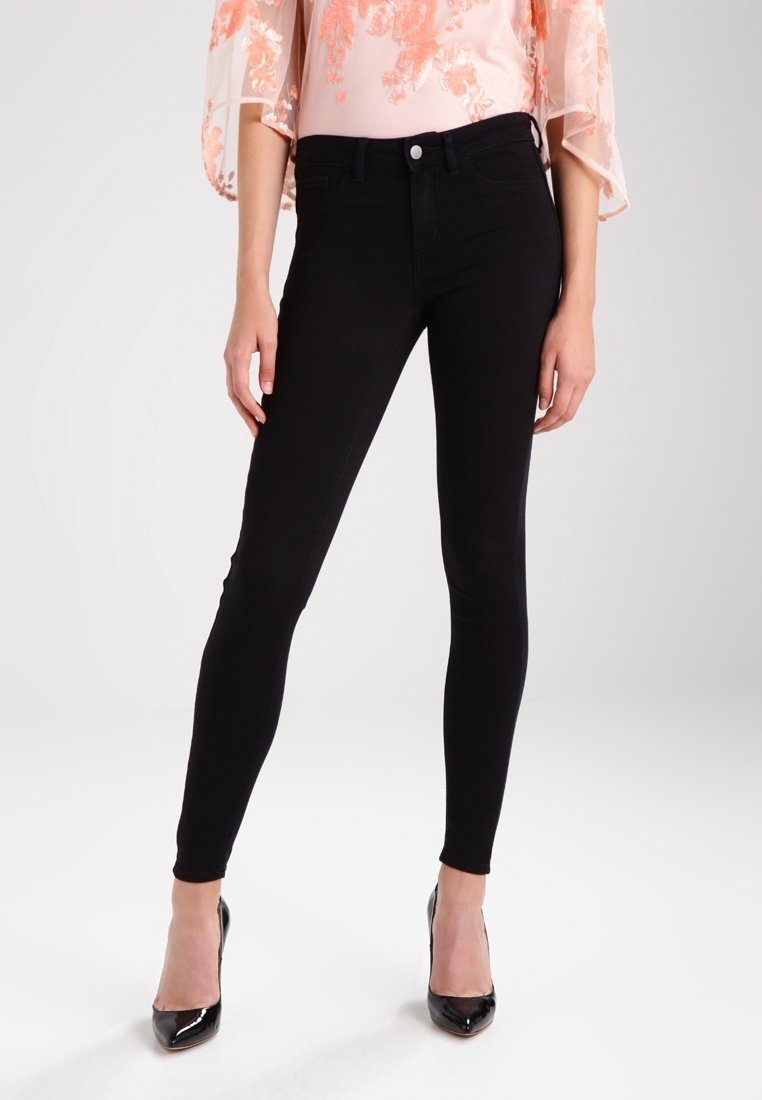 Femme portant un jean skinny noir taille haute associé à un haut en dentelle florale rose et des talons vernis noirs à bout pointu.