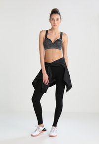 Fekete sportmelltartó texturált kivitelben, fekete leggings-szel és megkötött fekete dzsekivel párosítva. A szettet fehér és rózsaszín sportcipők egészítik ki.