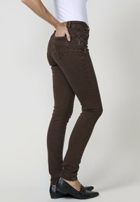 Jeans ajustados marrones de denim, con un diseño ceñido, bolsillos traseros con detalles y una textura suave. Combinados con zapatos negros.