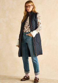 Zila kvadrātveida bezpiedurkņu veste ar brūnu apdari, uzlikta virs baltas rakstainas blūzes. Kombinēta ar ziliem džinsiem un brūnām mokasīnām, valkājot rozā punktiņu zeķes.