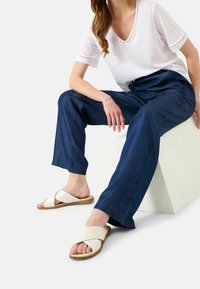 En lätt vit t-shirt med v-krage, marinblå byxor med snöre och beige sandaler med korskärning på en modell som sitter bredvid en geometrisk vit pelare.