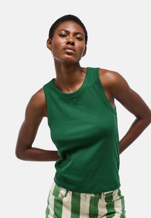 Jeune femme noire portant un haut vert sans manches et un pantalon rayé vert et blanc, posant avec les mains dans le dos.