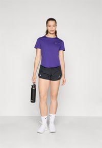 Ljubičasta Nike majica, crne Nike kratke hlače, bijele čarape i bijele tenisice. Drži crnu bocu za vodu, stoji na jednostavnoj pozadini.
