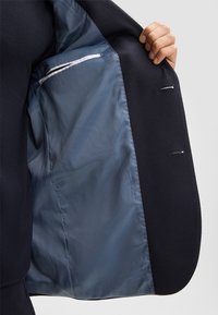 Blazer navy con un esterno testurizzato e una fodera liscia azzurro chiaro, caratterizzato da una tasca con bordatura bianca e due vani interni per accessori.