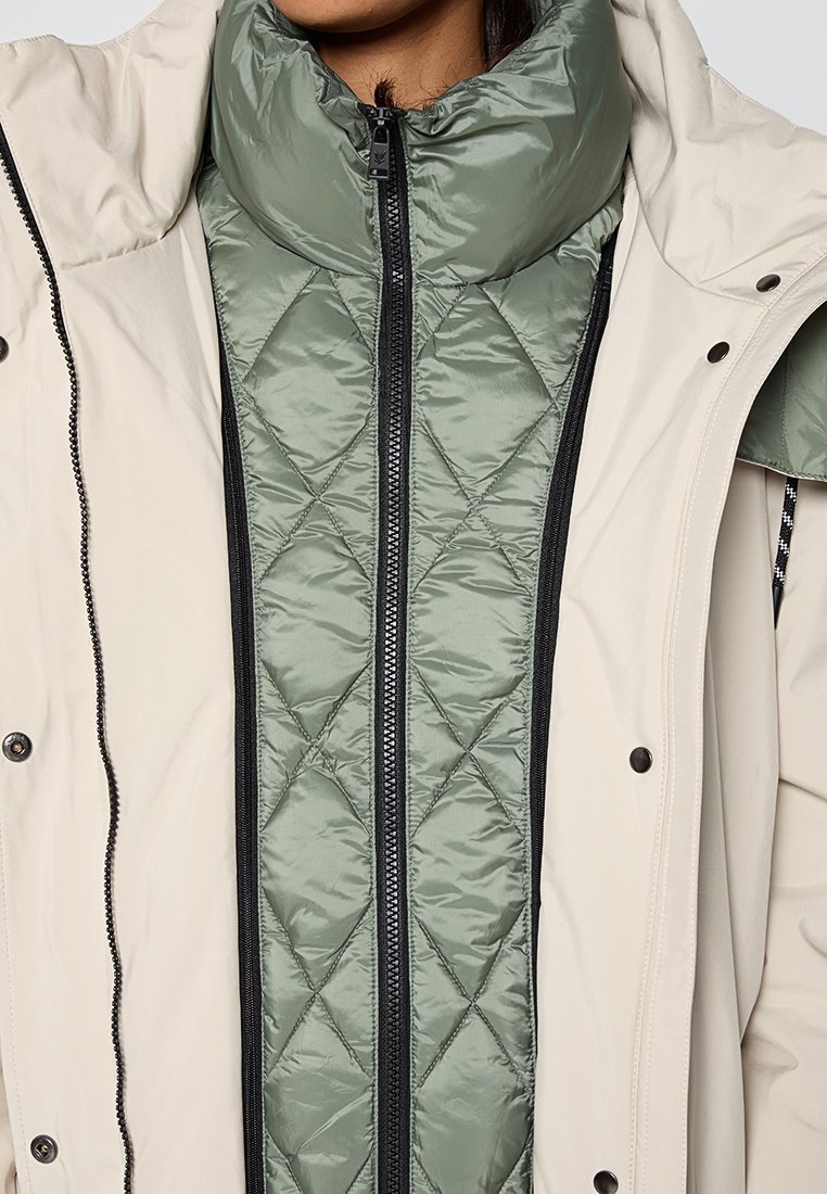 Personne portant des vestes matelassées à col haut, superposées en beige et vert, montrant des détails de texture du tissu et des fermetures.