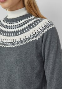 Woman wearing a grey sweater with white and black intricate geometric patterns around the yoke, layered over a white turtleneck.