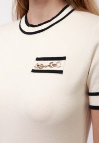 Crème korte mouwen top met een zwarte geribbelde kraag en mouwranden. Beschikt over een kleine zwarte zak met een zilveren kettingaccent.