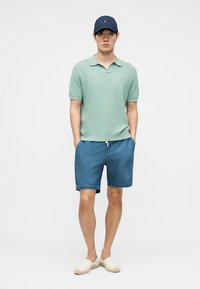 Mann iført en grønn kortermet polo, blå denimshorts, marineblå caps og beige slip-on sko, stående med hendene i lommer på ensfarget bakgrunn.