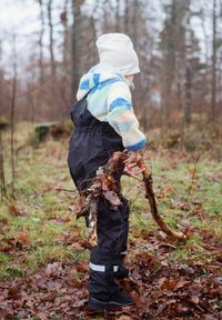 Svarta vattentäta overaller med reflexband, i kombination med en flerfärgad fleecejacka. Barnet håller i grenar omgiven av fallna löv.