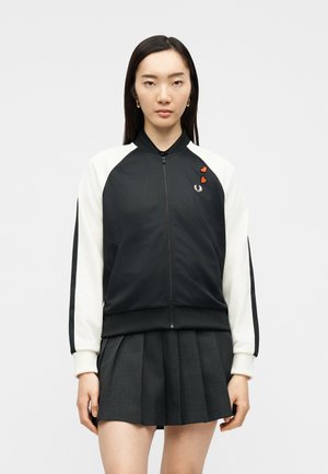 Nuori nainen, jolla on mustavalkoinen vetoketjullinen takki, jossa on kaksi punaista sydänmerkkiä, ja tumma laskostettu hame, katsoo suoraan eteenpäin.