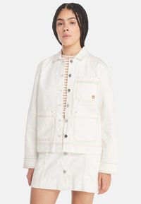 Hvit denimjakke med oransje sømmer, knappestolpe, to frontlommer og krage. Båret over en stripete skjorte.