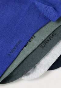 Keturių marškinėlių krūva: mėlyni, alyvuogių žalios, pilki ir juodi, pagaminti iš minkštos medvilnės su subtilia tekstūra, su juodais atspausdintais prekės ženklų ženklais.