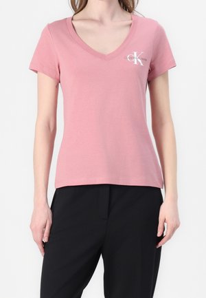 Kobieta ubrana w różowy t-shirt z krótkim rękawem i dekoltem w serek, z białym logo Calvin Klein, w połączeniu z czarnymi spodniami, stojąca na białym tle.