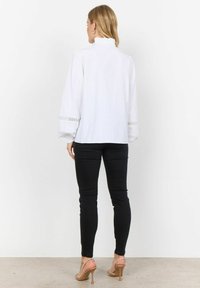 Blusa bianca con maniche lunghe voluminose, colletto alto arricciato e rifiniture in pizzo sulle maniche, abbinata a jeans skinny neri e tacchi nudi.