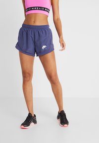 Kvinna iklädd marinblå Nike-shorts, rosa sportbehå och svarta sneakers med rosa sulor som står mot en enfärgad vit bakgrund.