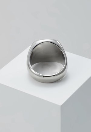 Anneau en métal argenté épais avec des bords lisses et arrondis, placé sur un socle blanc sur un fond gris clair uni.