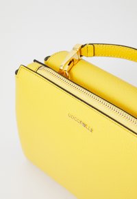 Borsa a mano gialla in pelle con una forma rettangolare strutturata, dotata di una cerniera dorata e di un manico ad anello. Accenti neri decorano i bordi.
